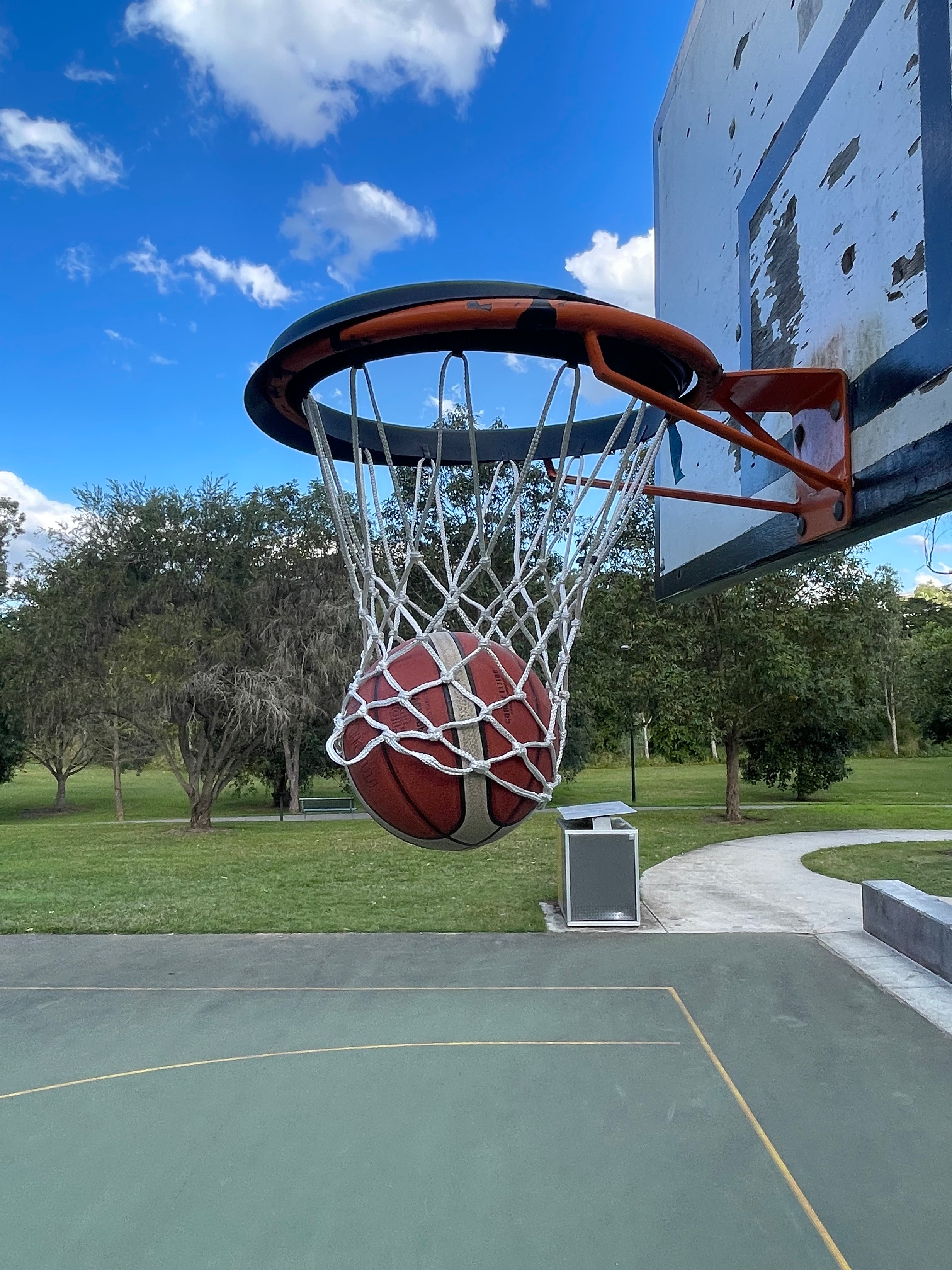 Basketball going through a hoop on an outdoor court with trees and blue sky in the background.